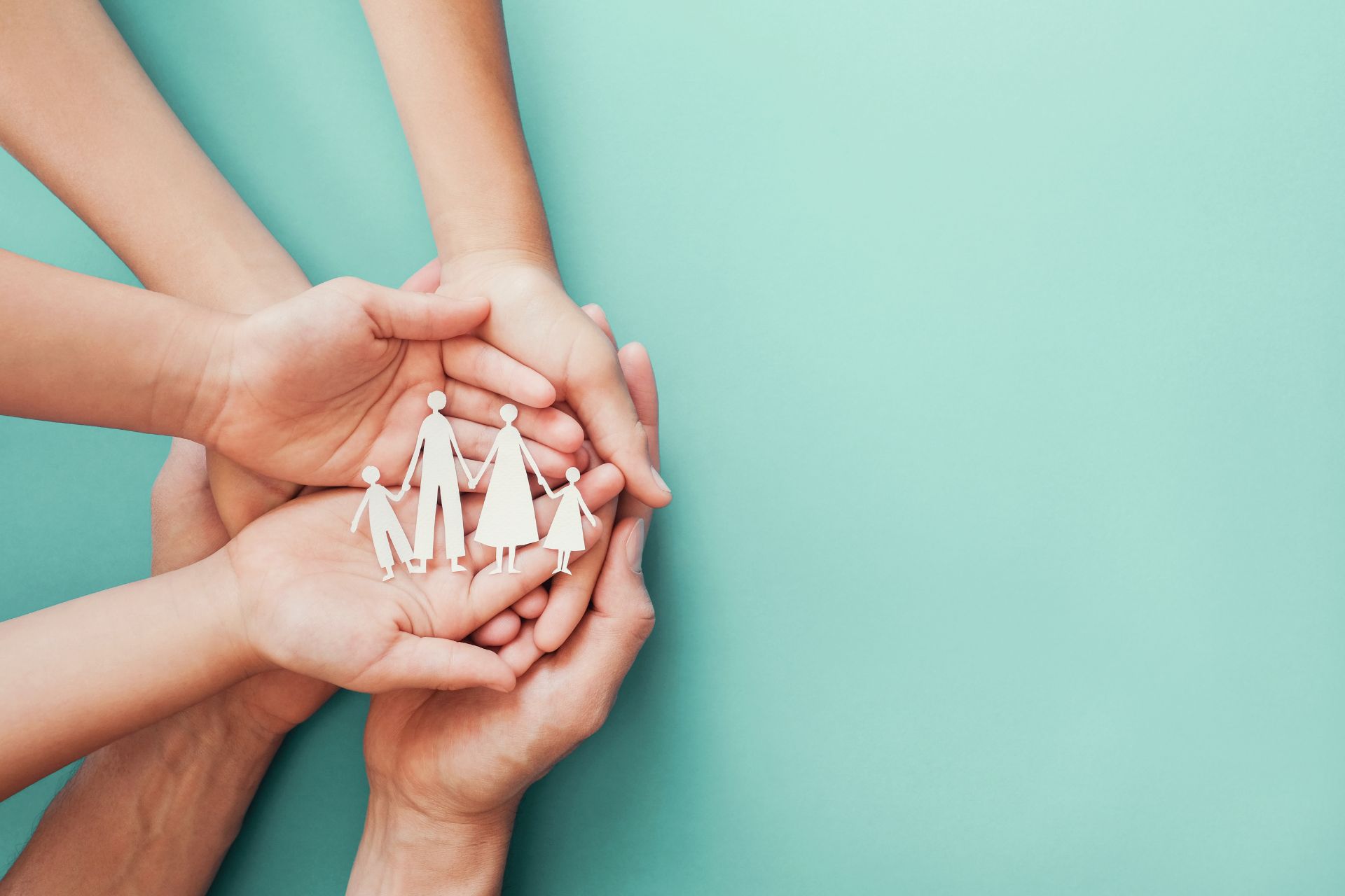 Draufsicht auf mehrere Hände von Erwachsenen und Kindern, die ineinandergelegt eine schützende Schale bilden. In der Mitte der Handflächen liegt eine kleine, aus weißem Papier ausgeschnittene Familie, bestehend aus zwei Erwachsenen und zwei Kindern, die sich an den Händen halten. Der Hintergrund ist in einem gleichmäßigen, matten Türkiston gehalten, mit viel freiem Raum auf der rechten Seite.